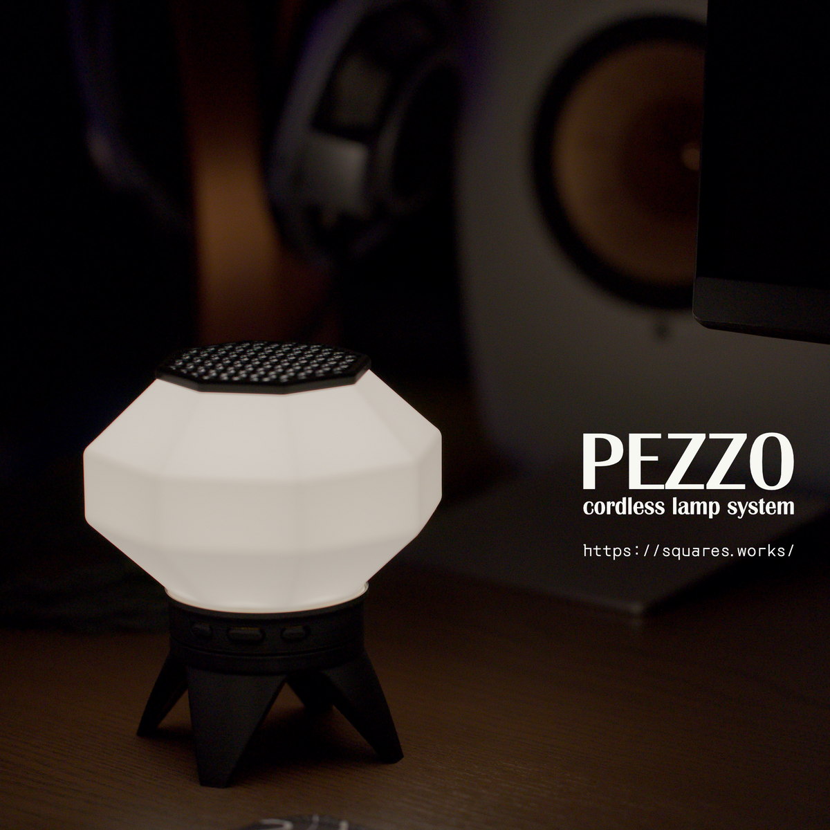 A modern white faceted cordless lamp with a black top grille and black tripod base, shown lit on a desk with a blurred computer monitor and audio equipment in the background. The text 'PEZZO cordless lamp system' is visible on the right.